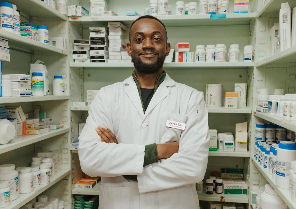 Pharmacist in a white coat stands with arms crossed in a pharmacy, surrounded by shelves of medicine bottles.