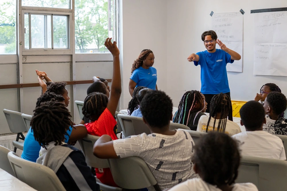 Two instructors engage with a group of children in a classroom; several children raise their hands.