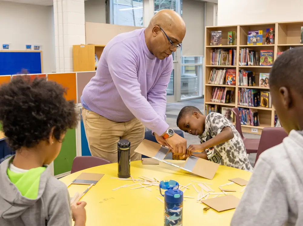 A teacher helps young students with a hands-on project at a table in a library or classroom.