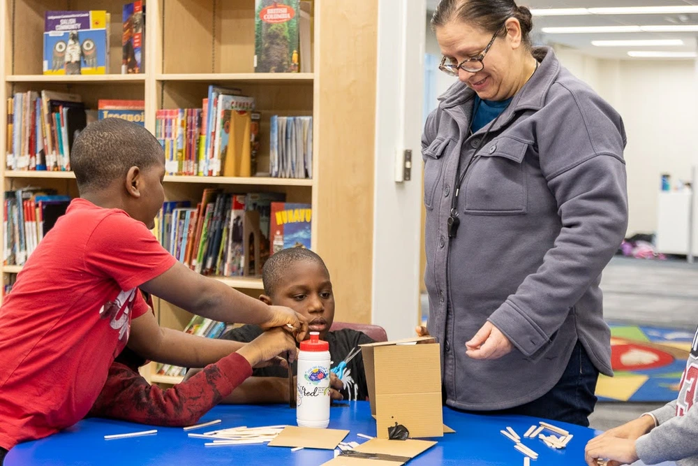 Three children and an adult work on a craft project at a table in a library, smiling and interacting.