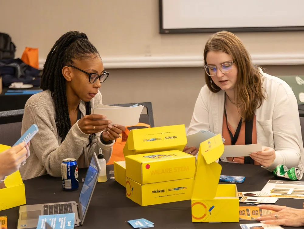 Two women sit at a table assembling STEM kits from yellow boxes, reading instructions and working together.