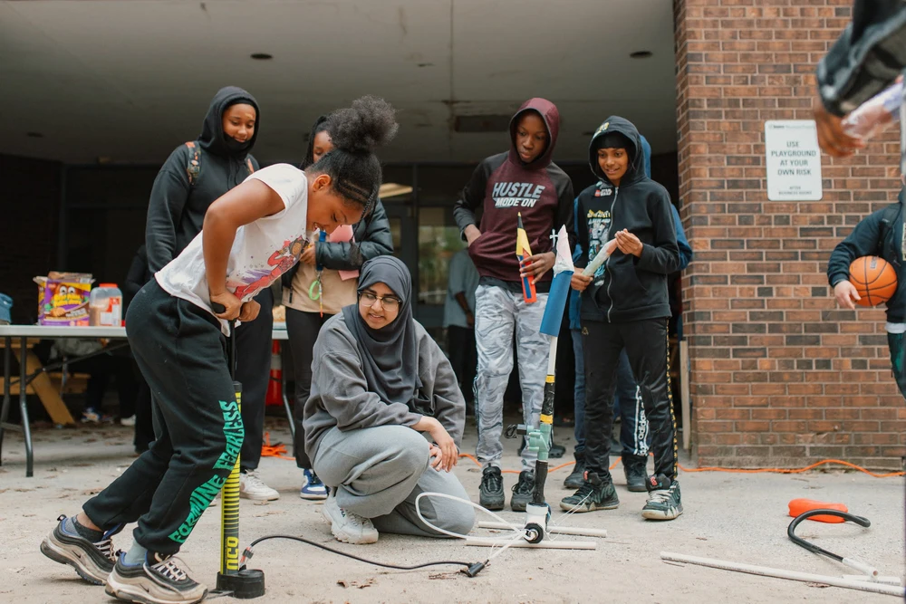 Children gather outside, launching model rockets, while one girl pumps air into a rocket launcher.