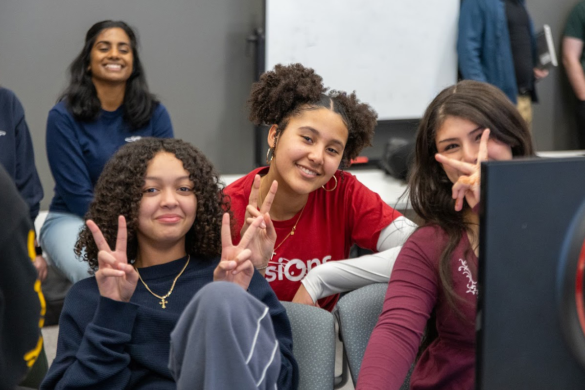 Three girls sitting and smiling, two making peace signs, with a woman smiling in the background.