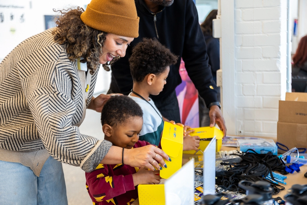 A woman and two children open yellow boxes at a table filled with gadgets and supplies.