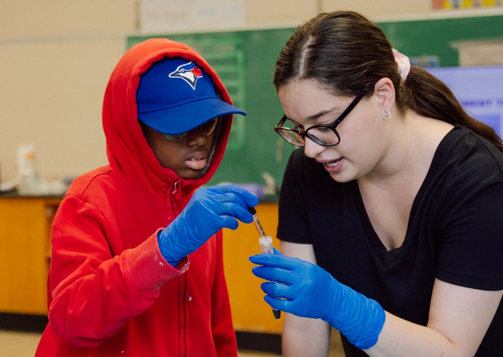A woman helps a boy in a red hoodie and blue cap with a science experiment in a classroom.
