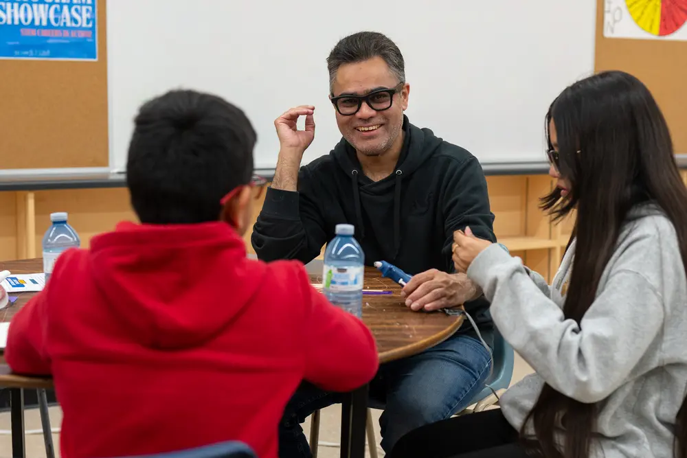 A man and two kids sit at a round table, smiling and talking in a classroom setting.