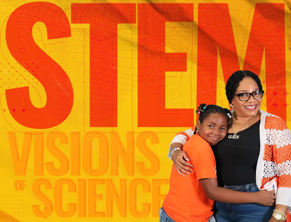 Two people smiling and hugging in front of a bright STEM VISIONS OF SCIENCE graphic background.