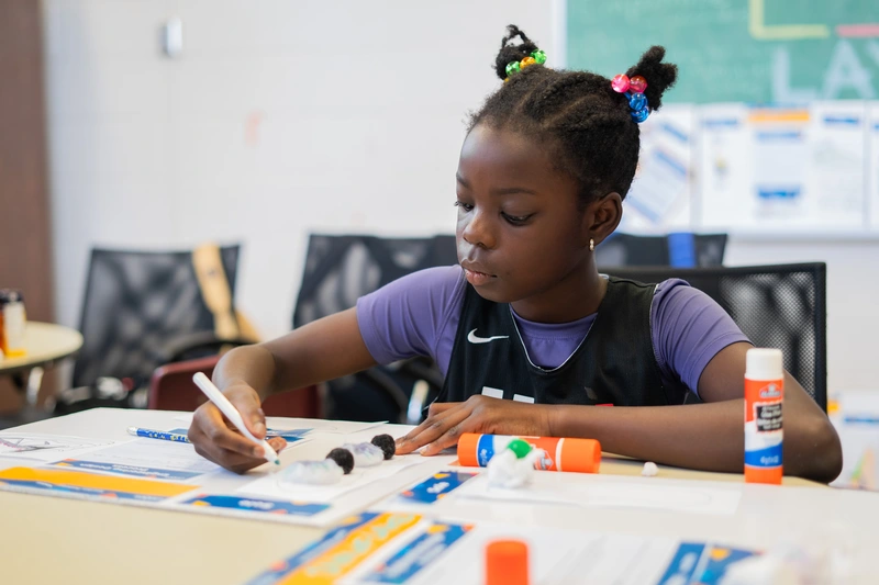 Young girl with colorful hair ties works on an arts and crafts project at a classroom table with glue sticks.