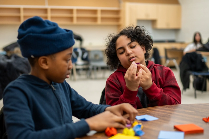 Two children sitting at a table, folding colorful origami paper in a classroom setting.