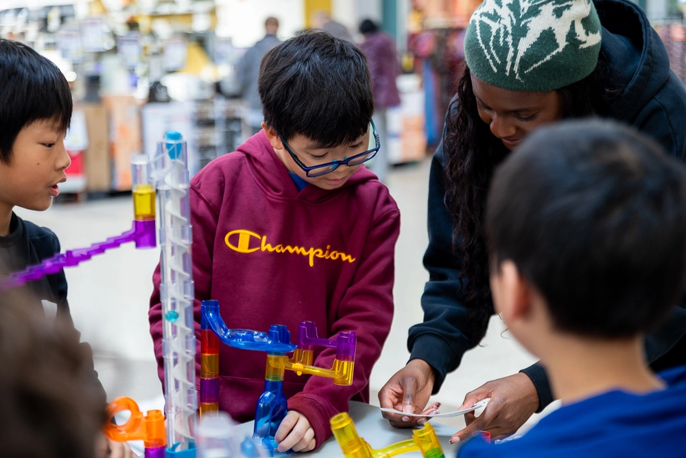 Four kids build a marble run together while one adult shows them instruction cards at a table.