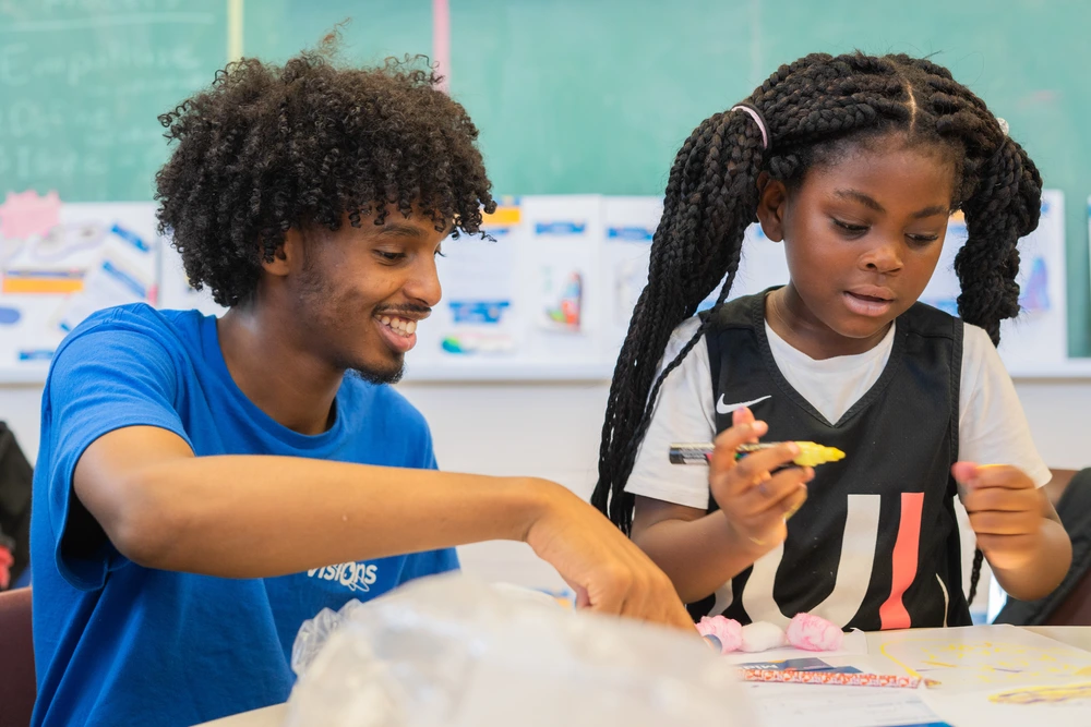 A man and a girl sit at a table doing crafts together in a classroom with a green chalkboard behind them.