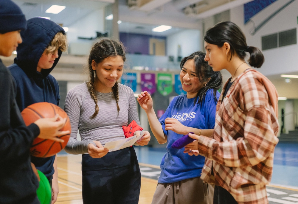 Four teens and an adult smile and talk together in a gym, holding papers, a gift, and a basketball.