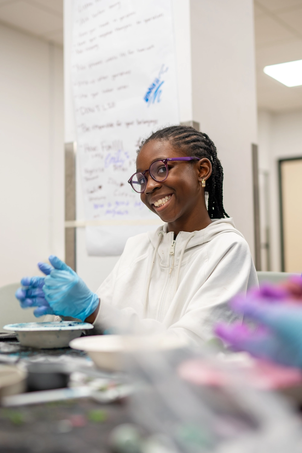 Smiling girl in glasses and gloves works on a project at a table in a classroom or lab setting.