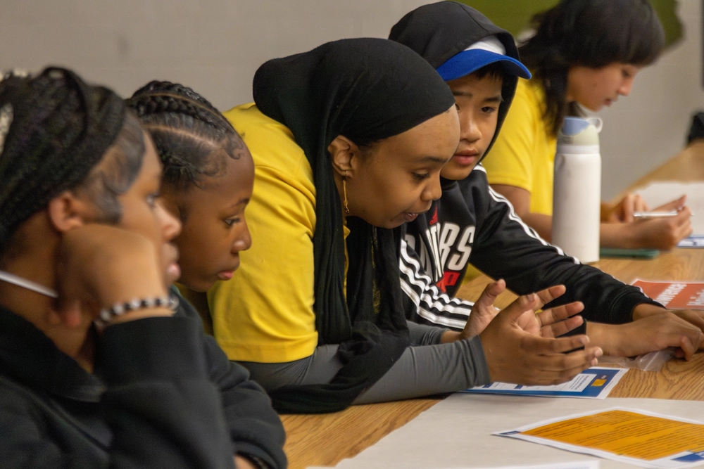 Four students sit at a table, focused on reading and discussing papers together in a classroom setting.