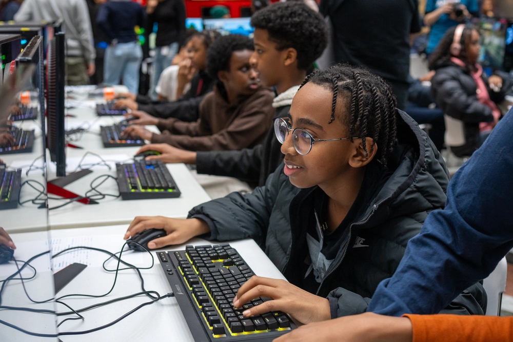 Children sit at computers, smiling and playing games together in a lively, indoor setting.