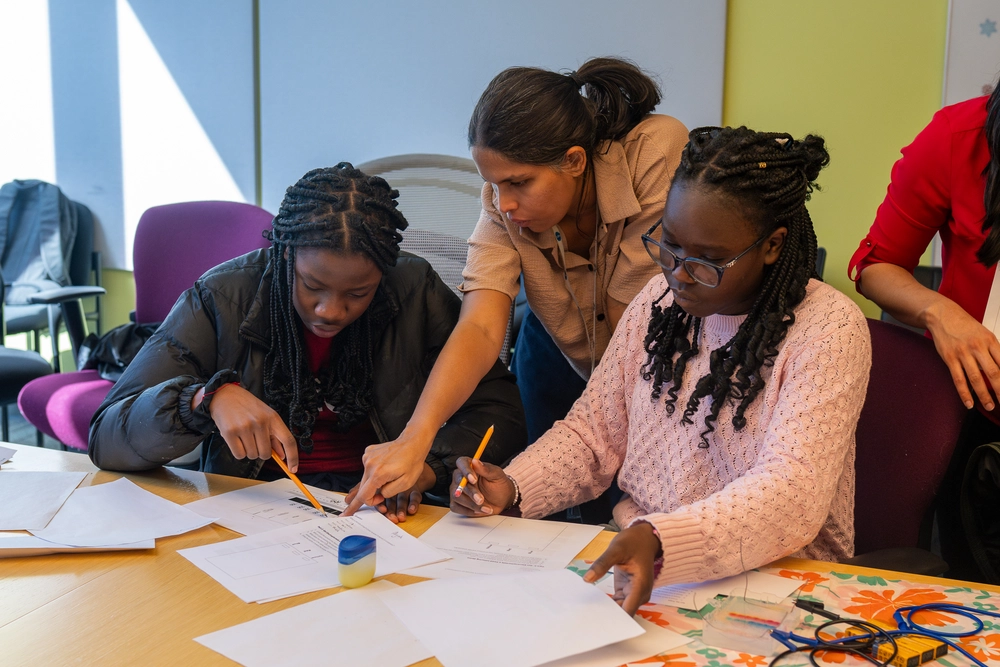 A teacher helps two students with worksheets at a table in a classroom setting.