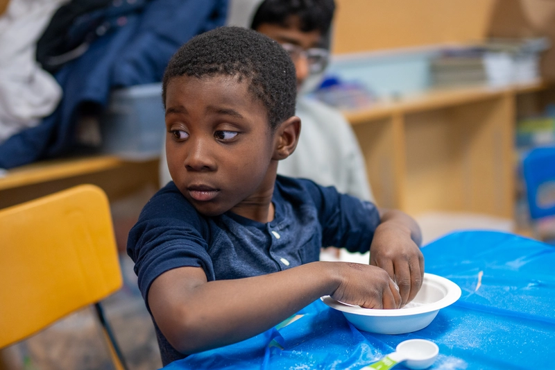 A young boy sits at a table, touching something in a bowl, and looks over his shoulder in a classroom.