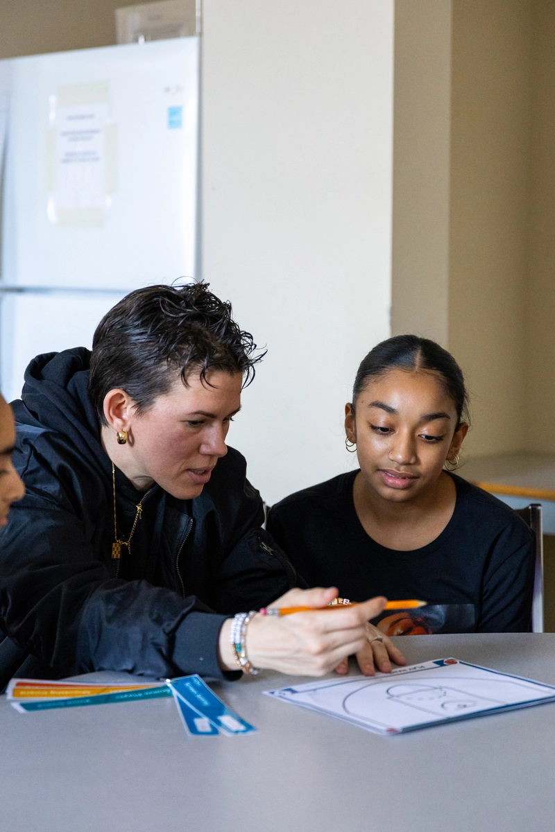 An adult helps a young girl with a worksheet at a table in a classroom setting.
