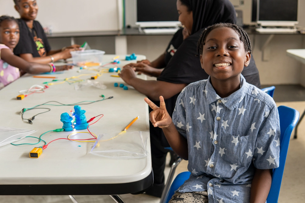 Smiling child flashes a peace sign at a table with electronics and craft materials; others work in the background.
