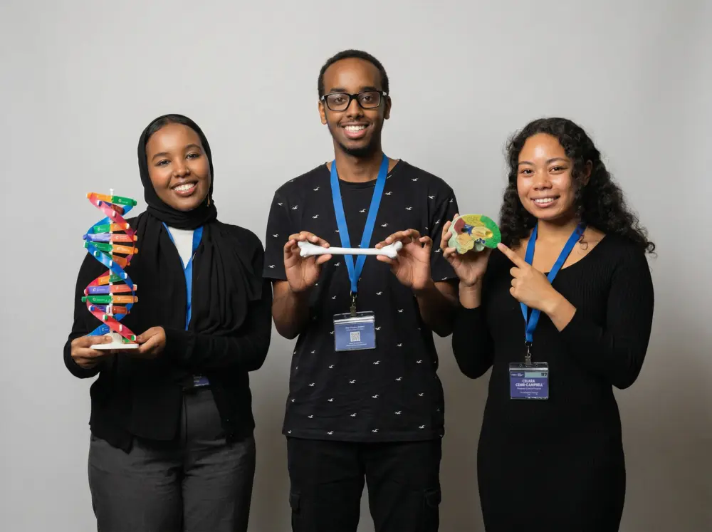 Three smiling students with lanyards hold science models: a DNA helix, a bone, and a model brain.