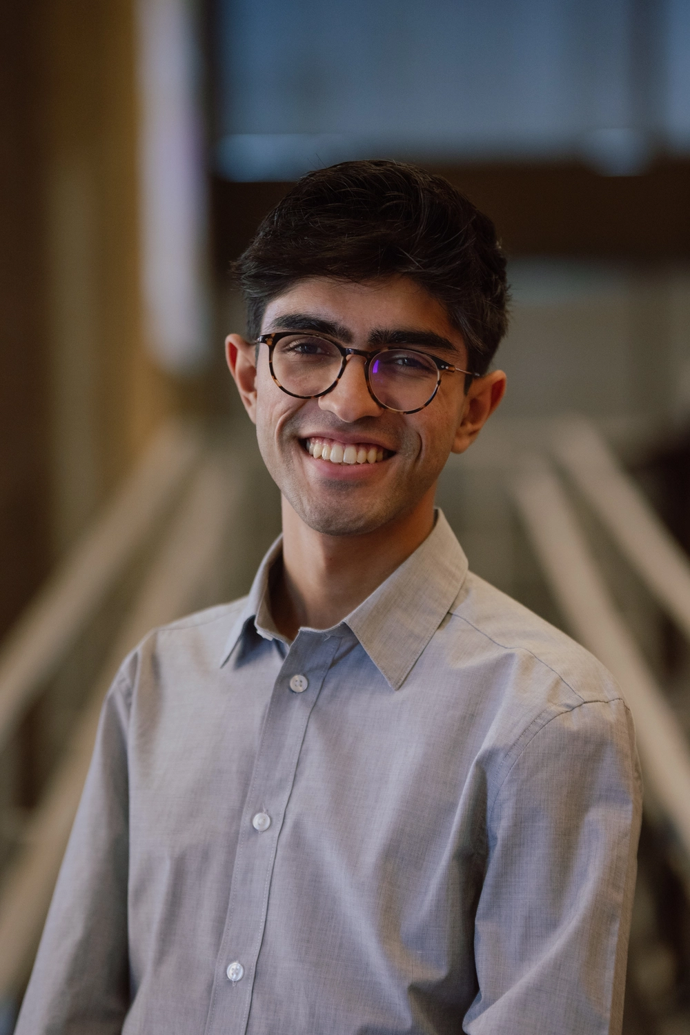 Young man with glasses smiling, wearing a light gray button-up shirt, standing indoors with blurred background.