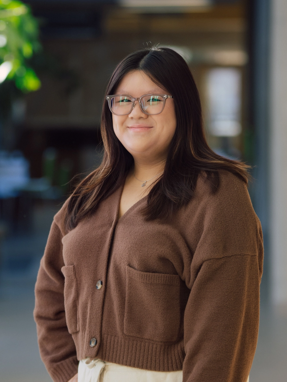 A woman with long dark hair and glasses smiles while wearing a brown cardigan, standing indoors.