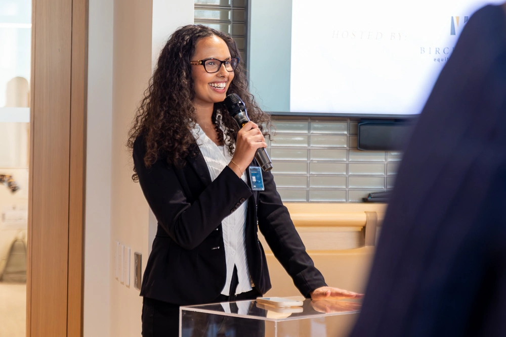 A woman in business attire smiles while speaking into a microphone at a podium indoors.