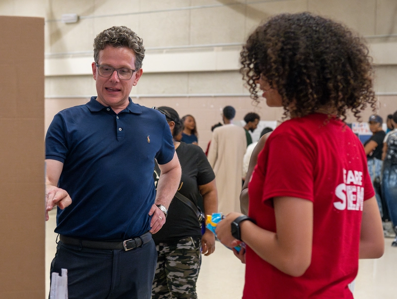 A man in a blue shirt talks to a girl in a red shirt at an indoor event with other people in the background.