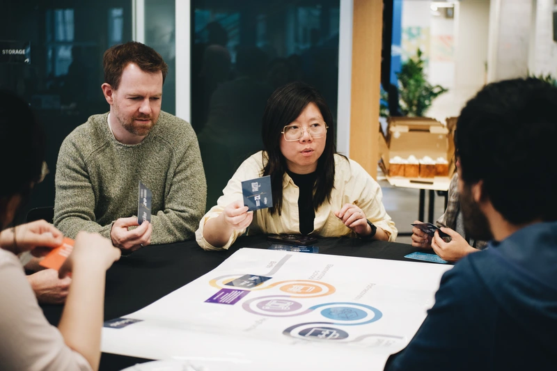 A group of people sit around a table discussing cards and a large chart during a collaborative meeting.