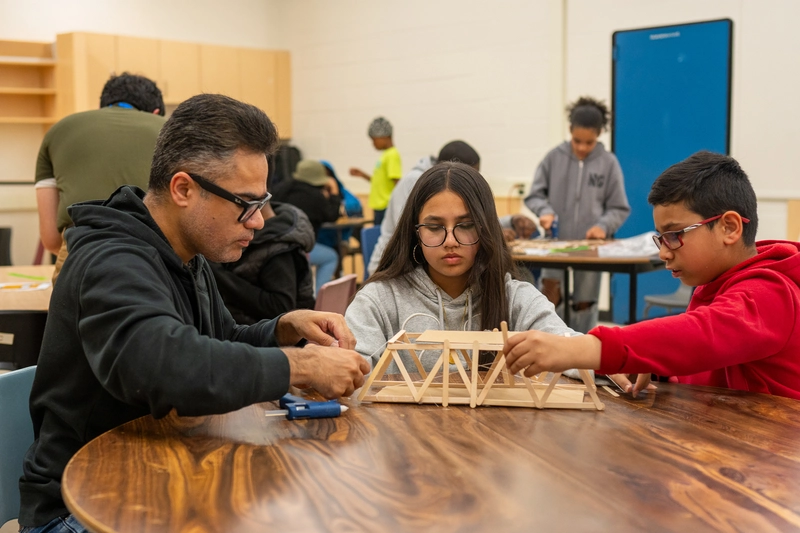 A teacher helps two students build a model bridge with wooden sticks in a classroom.