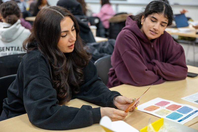 Two students sitting at a desk, discussing a card and a colorful worksheet in a classroom setting.