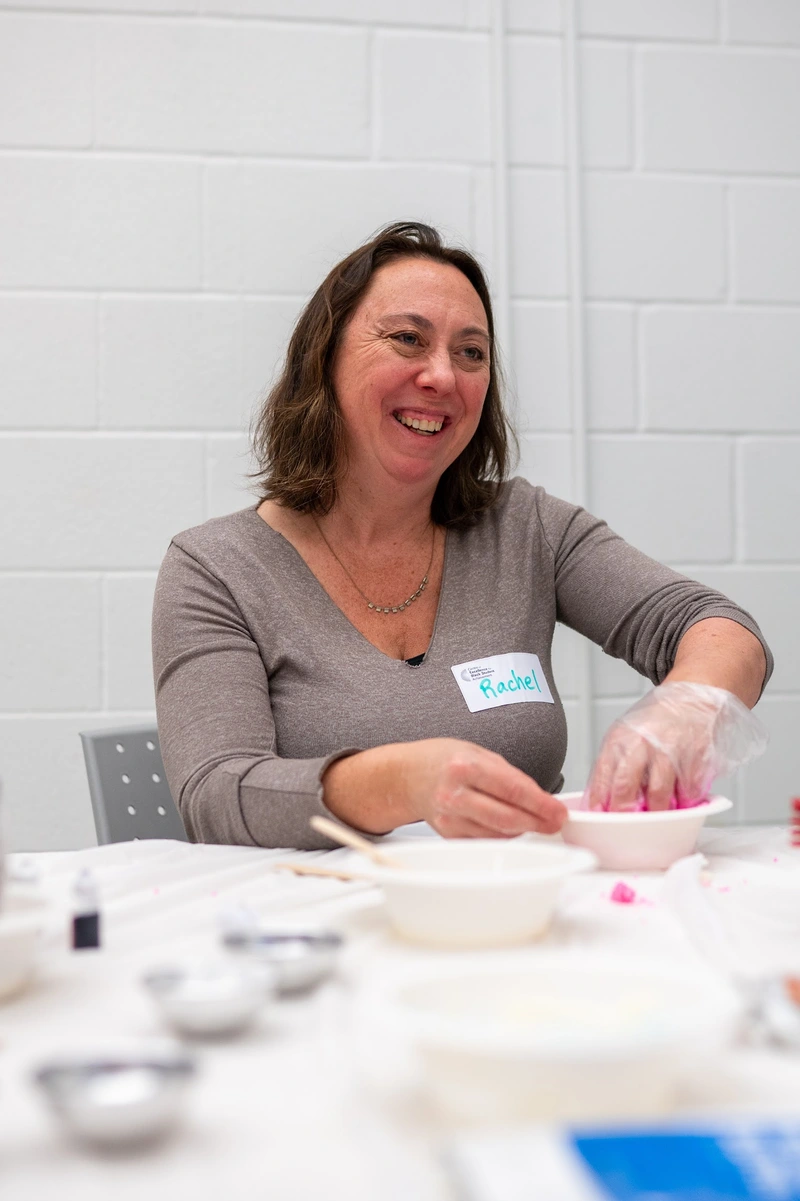 Smiling woman wearing a name tag sits at a table, mixing something in a bowl with gloved hands.
