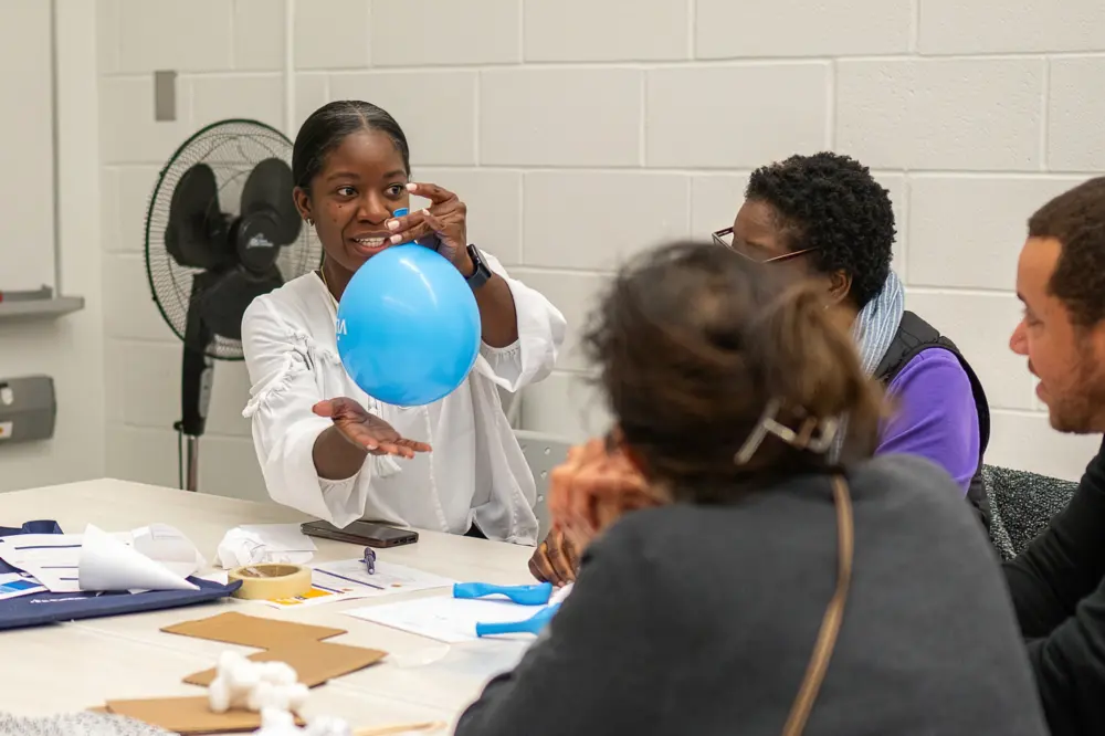 A woman holds a blue balloon, smiling, while seated with three others at a table in a classroom setting.