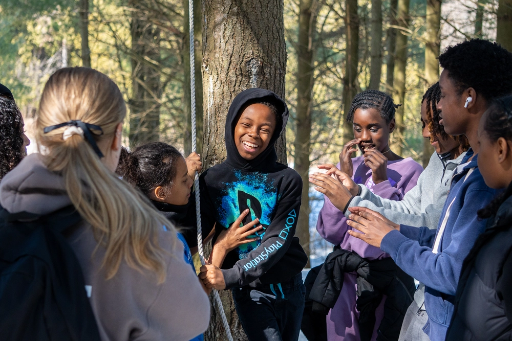 A group of kids laughing and talking together around a tree in a sunlit forest.
