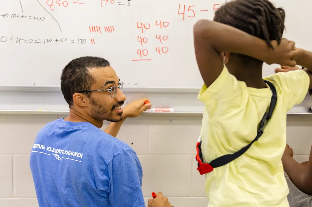 A teacher writes math problems on a whiteboard while a student stands nearby, watching attentively.