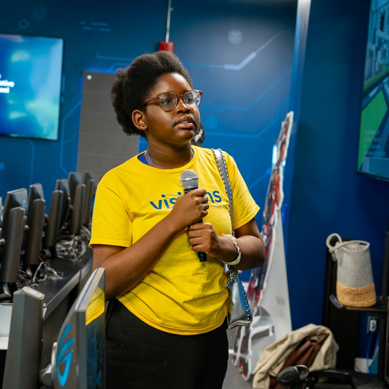 A young person in a yellow visitors shirt holds a microphone in a modern computer lab.