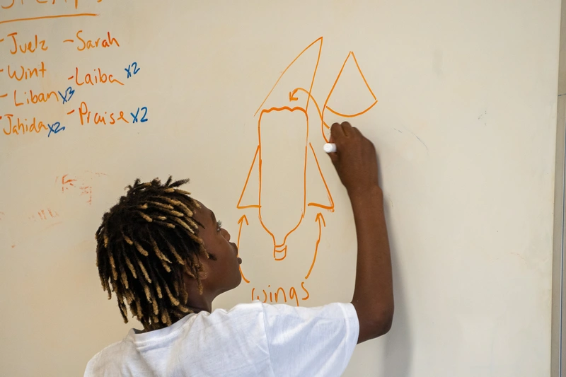 A child draws a rocket diagram labeled Wings on a whiteboard with names written at the side.