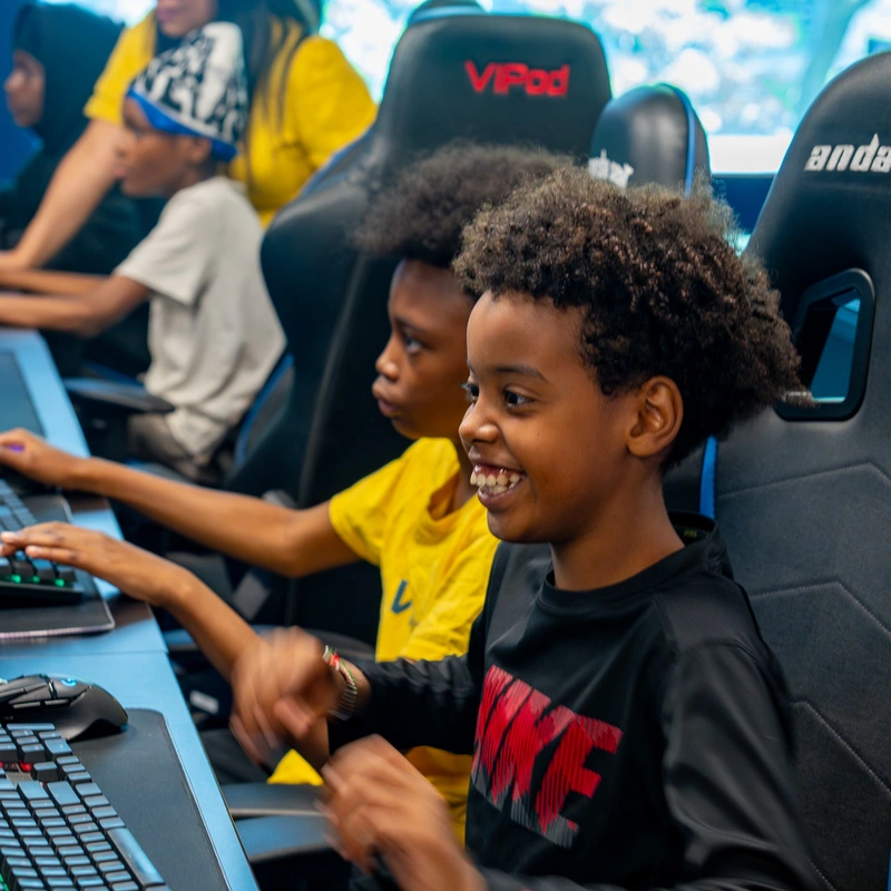 Smiling kids playing on computers in a gaming room, sitting in large black gaming chairs.