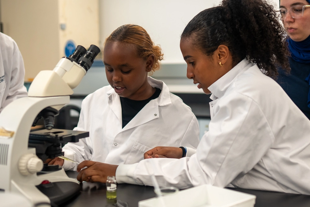 Two students in lab coats work together at a microscope in a science laboratory.