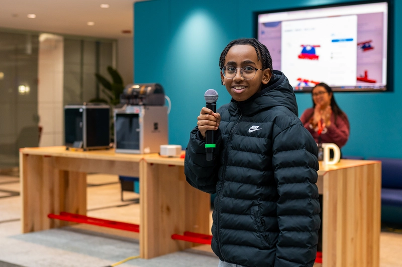 A smiling young person in a black jacket holds a microphone in a modern room with a screen and wooden table.