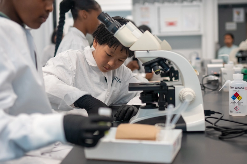 Children in lab coats examine samples with a microscope in a science classroom or laboratory setting.