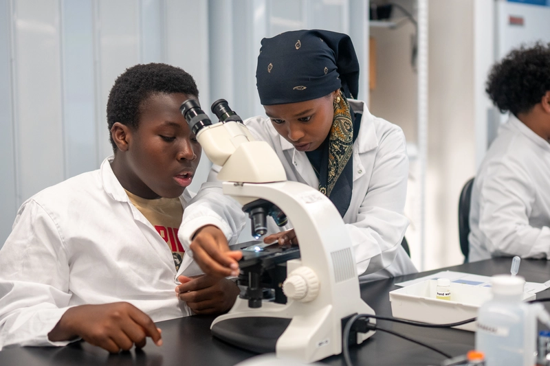 Two students in lab coats use a microscope together in a science classroom.