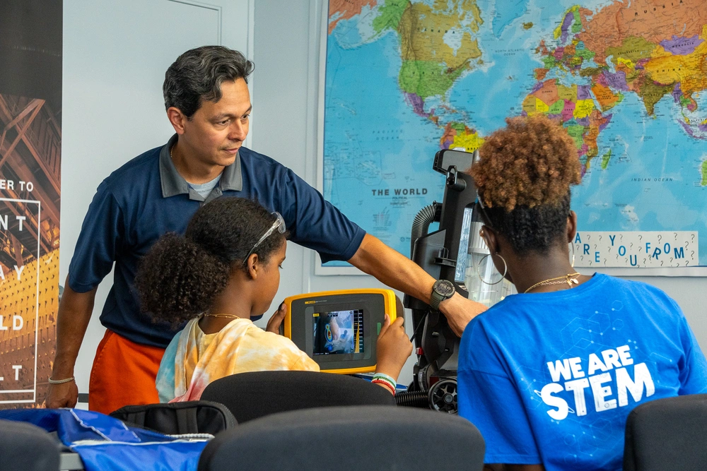 A teacher helps two students use scientific equipment in a classroom with a world map on the wall.