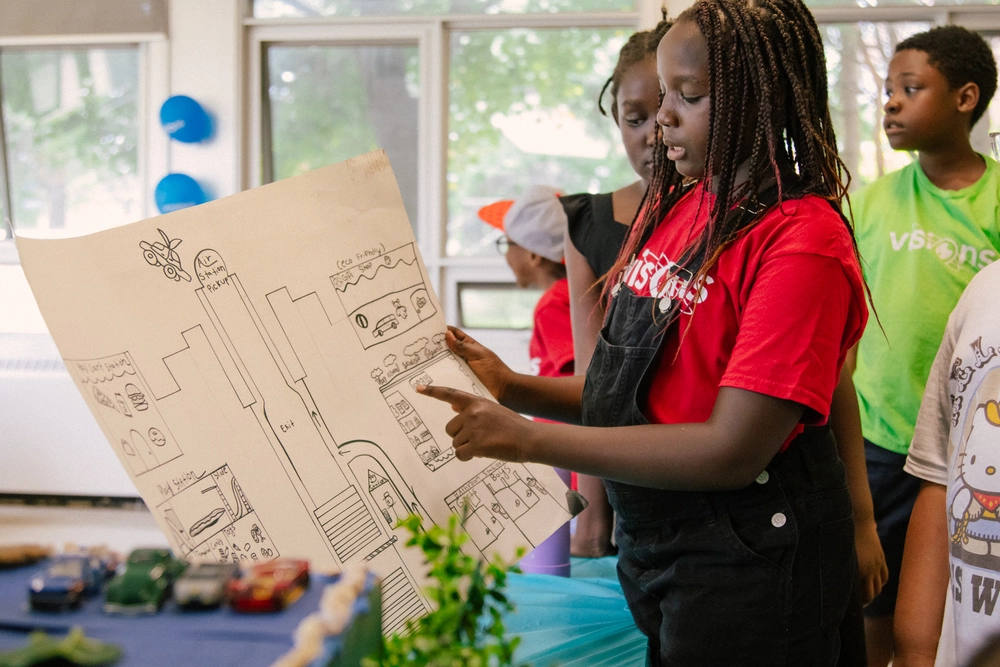 A girl shows a large hand-drawn city map to a group of children in a classroom.