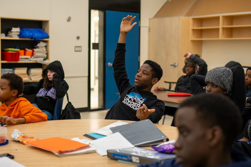 A student raises his hand in a classroom while others sit and listen around a round table.