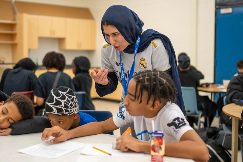 A teacher helps two boys with schoolwork at a classroom table; one boy is wearing a spiderweb hat.