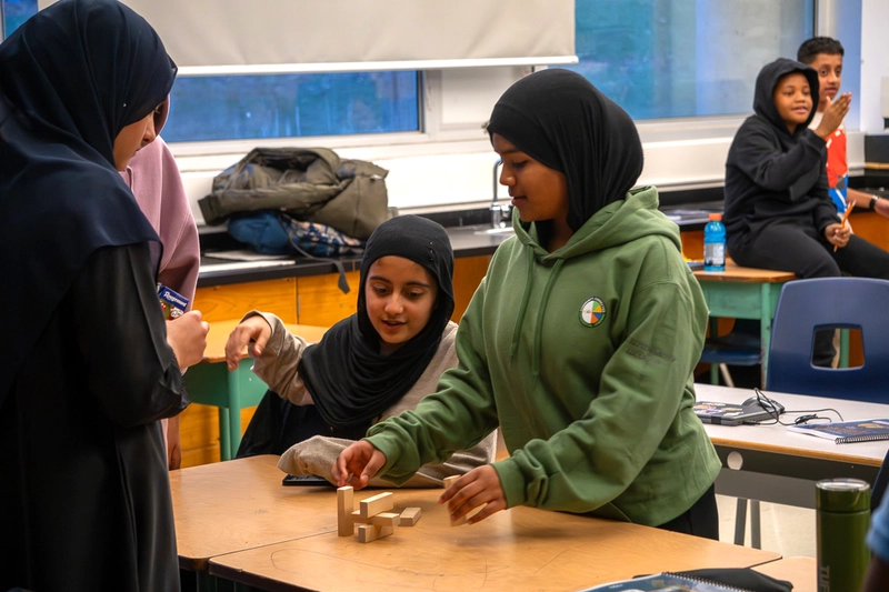 Three girls in hijabs work together to stack wooden blocks in a classroom; other students are in the background.