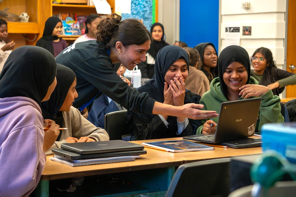 A group of students smile and look at a laptop together in a lively classroom setting.
