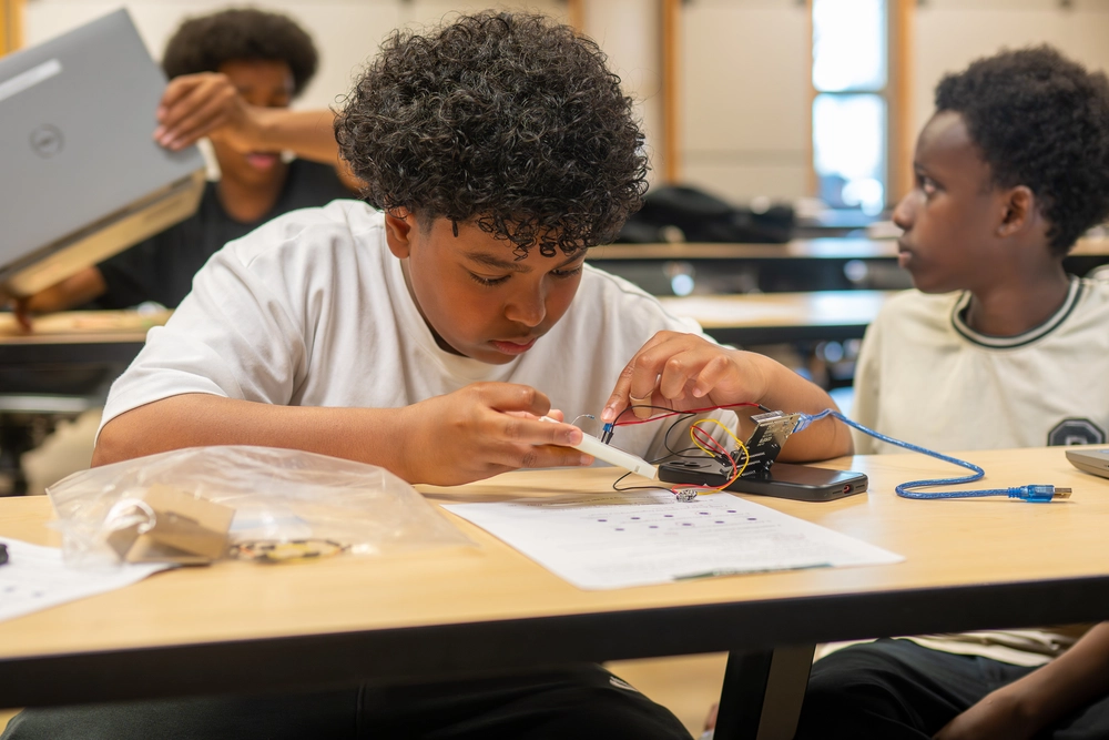 Boy focuses on assembling an electronic circuit at a classroom desk, with two other boys nearby.