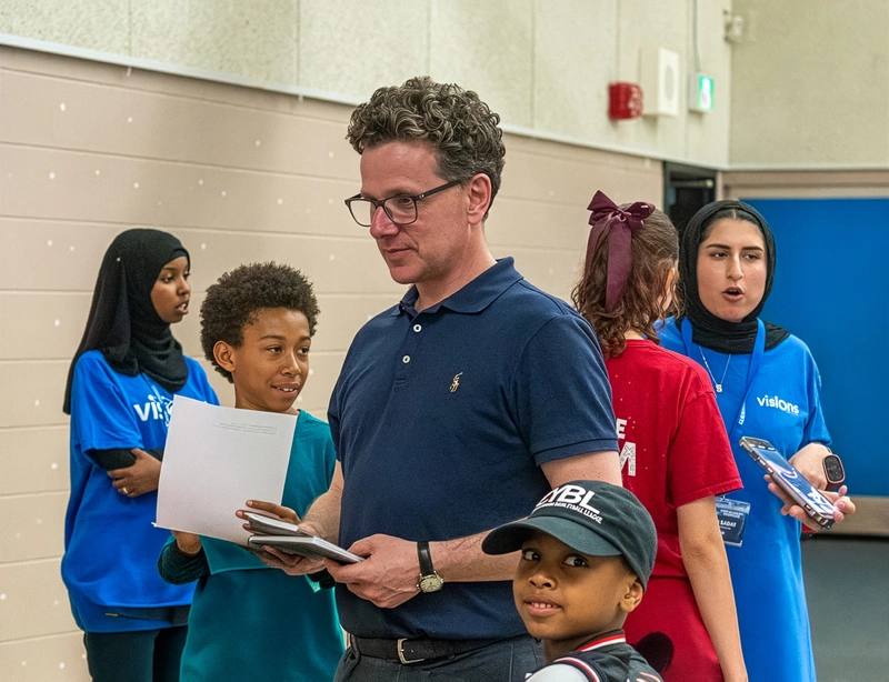 A group of people, adults and children, stand indoors holding papers and talking at an event.